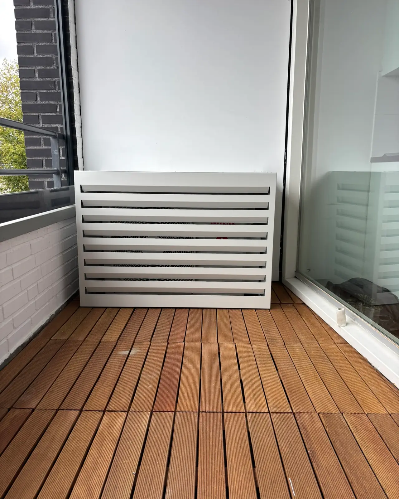 Een buitendeel in een witte lamellenkast op het balkon van een appartement, op een houten vloer.