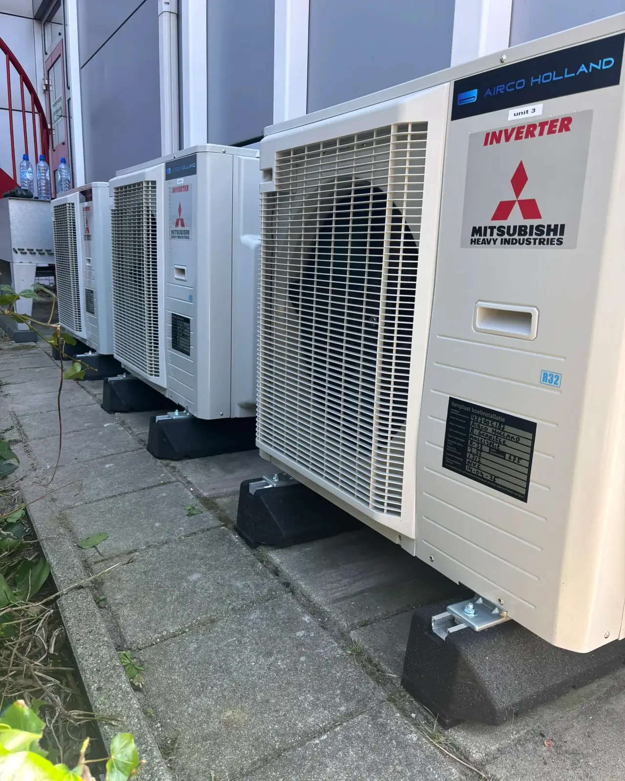 Een rij buitendelen van Mitsubishi Heavy Industries op voetjes langs de gevel van een bedrijfspand.